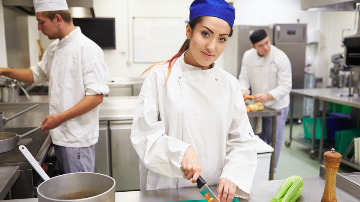 Drei junge Menschen arbeiten in einer Küche. Im Vordergrund steht eine junge Frau, die Gemüse schneidet.