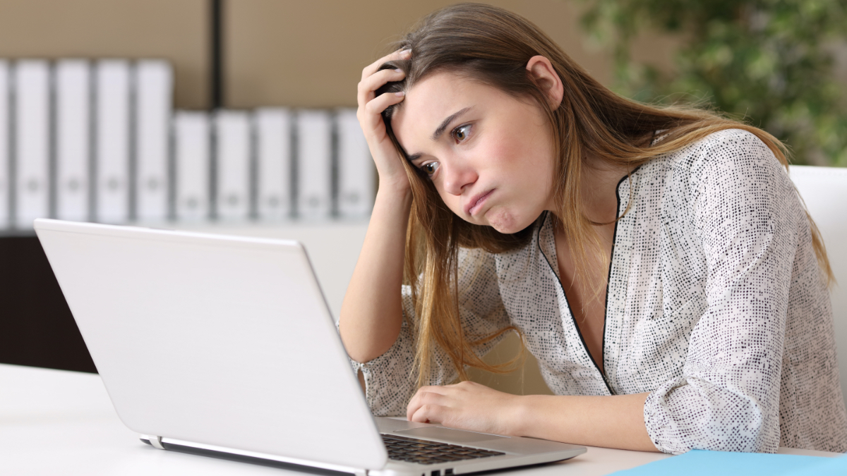 Eine junge Frau mit verzweifeltem Gesichtsausdruck sitzt vor einem Laptop und stützt ihren Kopf auf einer Hand ab.