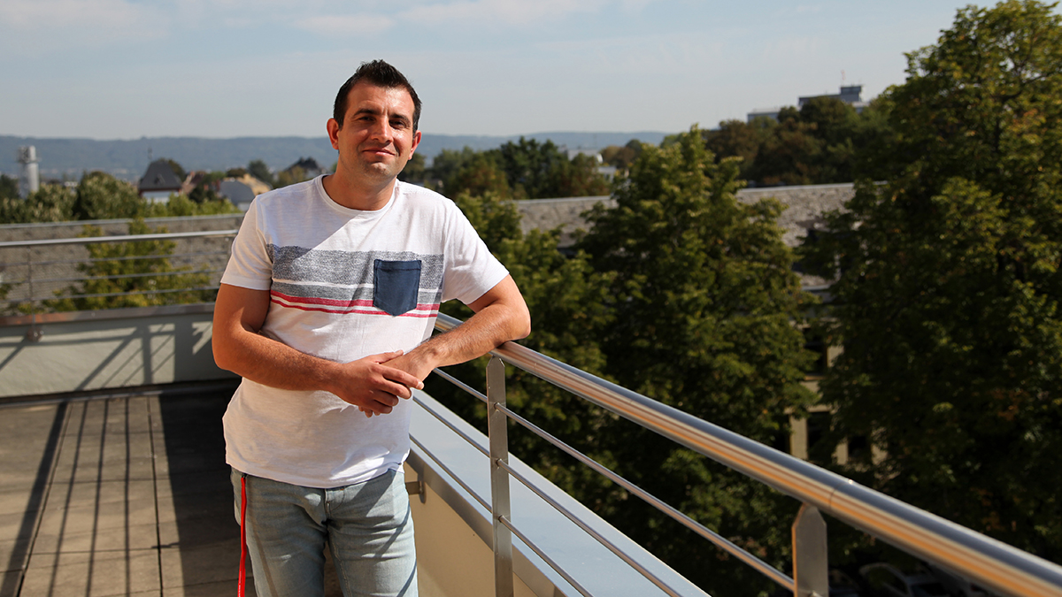 Ein Mann steht auf einer Dachterrasse und lehnt sich mit einem Arm auf das Geländer. (Quelle: BAMF) Ein Mann steht auf einer Dachterrasse und lehnt sich mit einem Arm auf das Geländer.