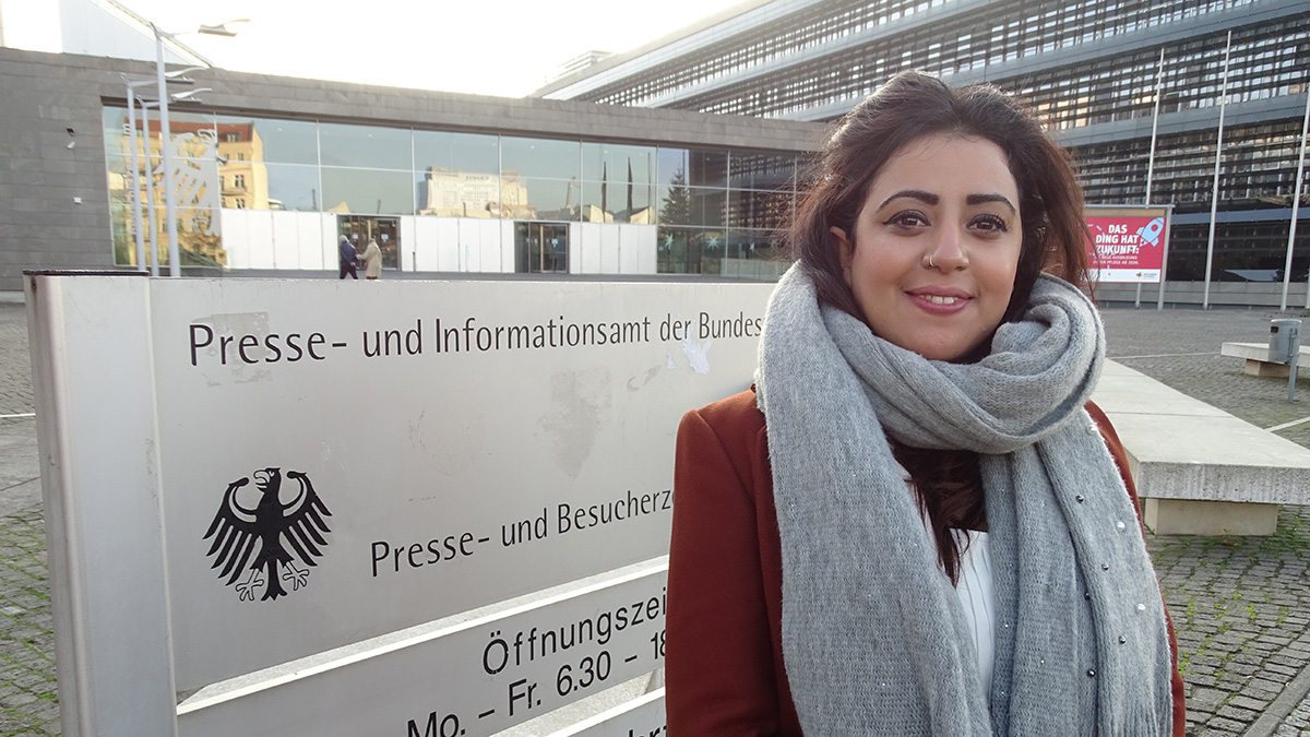 Eine junge Frau steht vor einem Schild am Eingang eines Gebäudes. (Quelle: BAMF | Tomšić) Eine junge Frau steht vor einem Schild am Eingang eines Gebäudes.