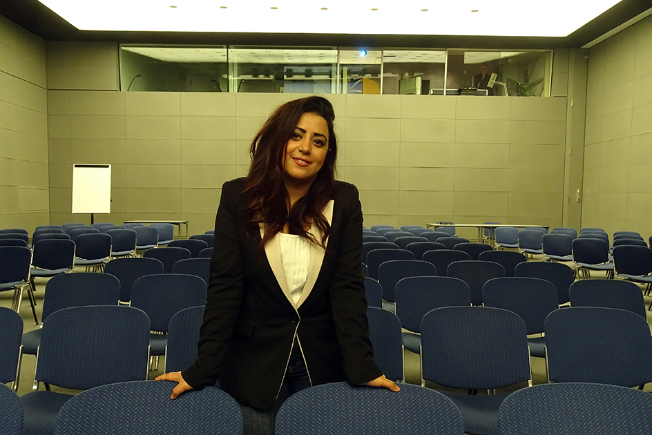 Eine junge Frau steht in einer Stuhlreihe in einem Saal. (Quelle: BAMF | Tomšić) Eine junge Frau steht in einer Stuhlreihe in einem Saal.