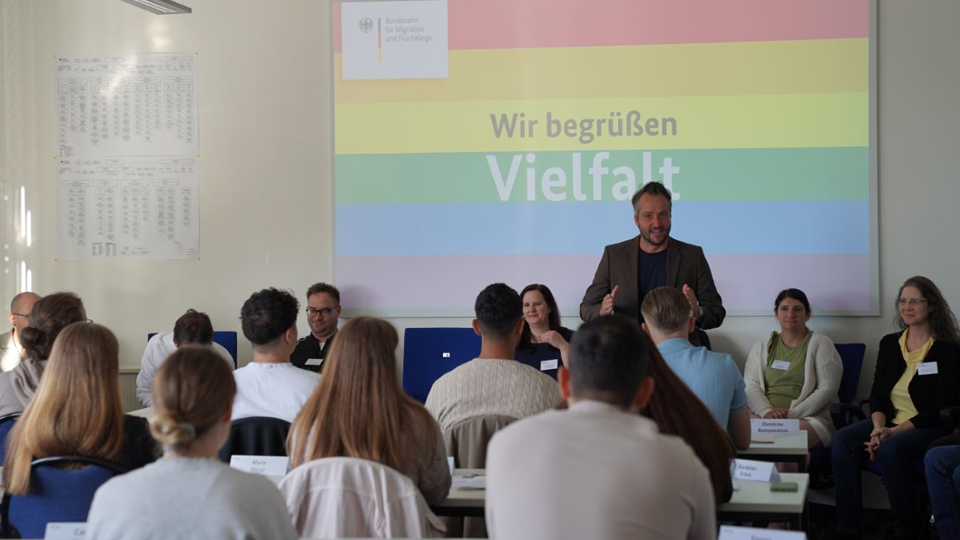 Im Vordergrund sitzen Menschen, die man von hinten sieht. In der Mitte steht ein Mann und redet; im Hintergrund der Schriftzug "Wir begrüßen Vielfalt" auf Regenbogenhintergrund. (Quelle: BAMF) Im Vordergrund sitzen Menschen, die man von hinten sieht. In der Mitte steht ein Mann und redet; im Hintergrund der Schriftzug "Wir begrüßen Vielfalt" auf Regenbogenhintergrund.