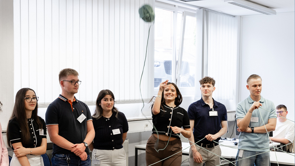 Sechs junge Menschen stehen in einem Halbkreis. Eine Person wirft ein Wollknäuel, zwei andere halten einen Teil der Schnur. (Quelle: BAMF) Sechs junge Menschen stehen in einem Halbkreis. Eine Person wirft ein Wollknäuel, zwei andere halten einen Teil der Schnur.