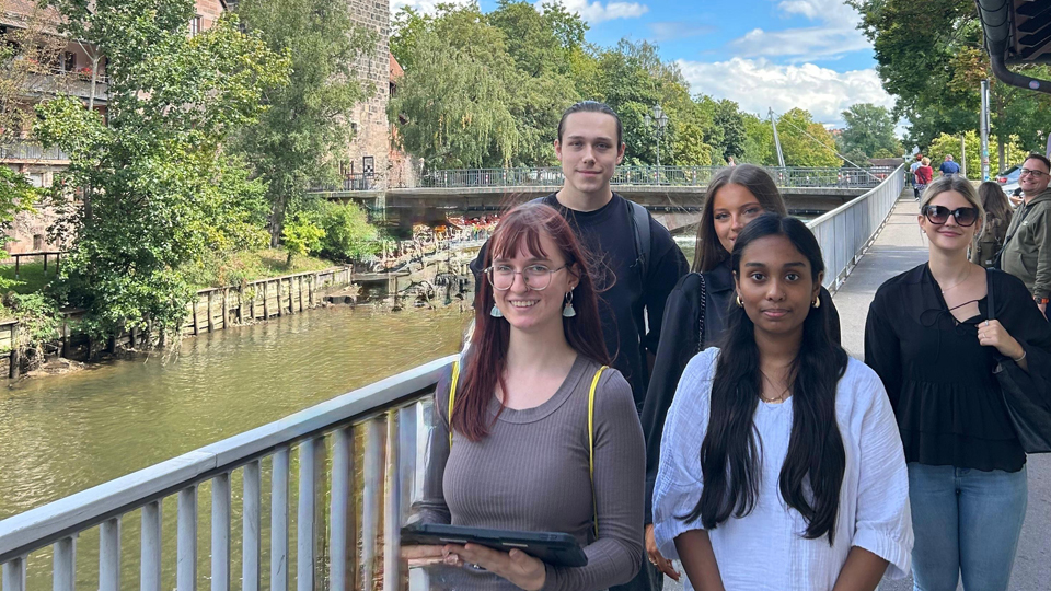 Fünf junge Menschen stehen an einem Geländer. Im Hintergrund ist ein Fluss und ein altes Gebäude. (Quelle: BAMF) Fünf junge Menschen stehen an einem Geländer. Im Hintergrund ist ein Fluss und ein altes Gebäude.