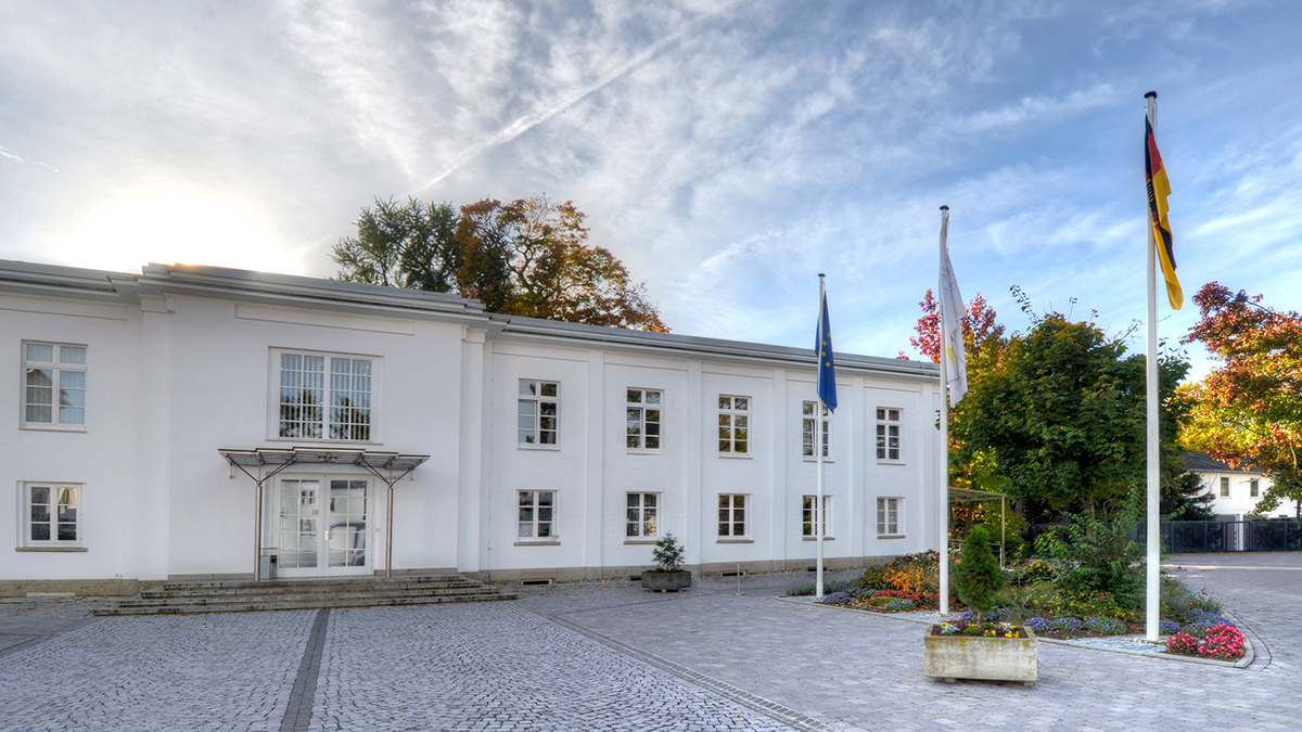 Auf einem Platz stehen drei Flaggen vor einem weißen Gebäude. (Quelle: Bundeskartellamt) Auf einem Platz stehen drei Flaggen vor einem weißen Gebäude.