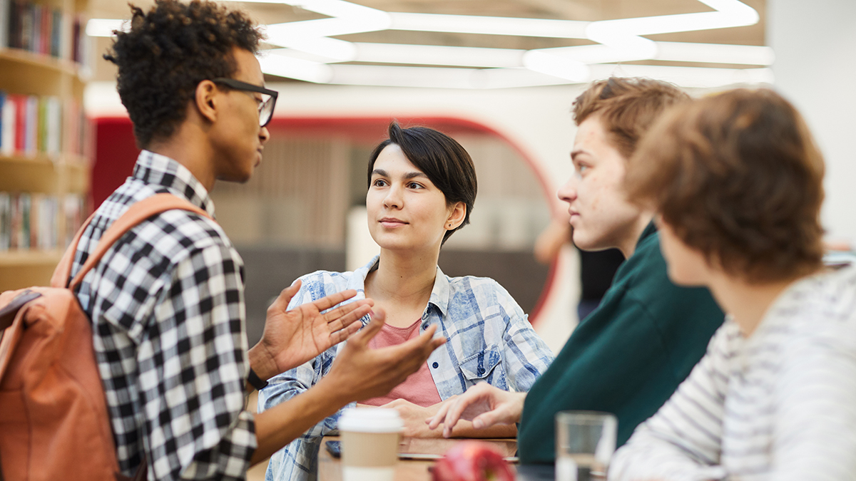 Vier junge Menschen stehen an einem Tisch und unterhalten sich. (Quelle: stock.adobe.com | Seventyfour) Vier junge Menschen stehen an einem Tisch und unterhalten sich.