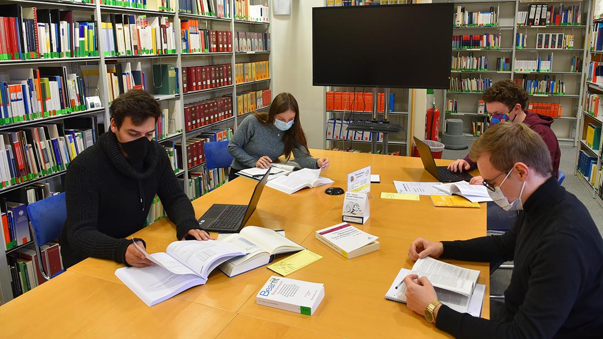 Vier junge Menschen sitzen mit Abstand und Masken an einem großen Tisch in einer Bibliothek und arbeiten mit Büchern. (Quelle: HS Bund) Vier junge Menschen sitzen mit Abstand und Masken an einem großen Tisch in einer Bibliothek und arbeiten mit Büchern.