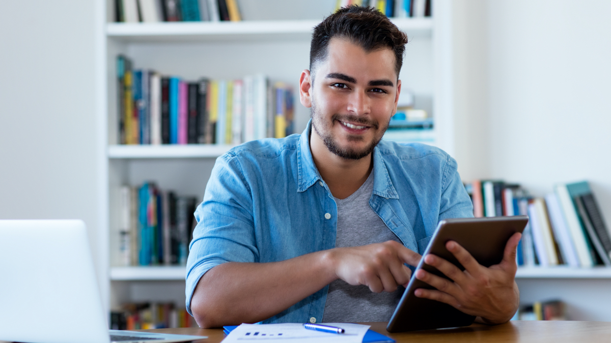 Ein junger Mann mit dunklen Haaren und Bart sitzt an einem Schreibtisch, lächelt und bedient ein Tablet. (Quelle: iStock | DMEPhotography) Ein junger Mann mit dunklen Haaren und Bart sitzt an einem Schreibtisch, lächelt und bedient ein Tablet.