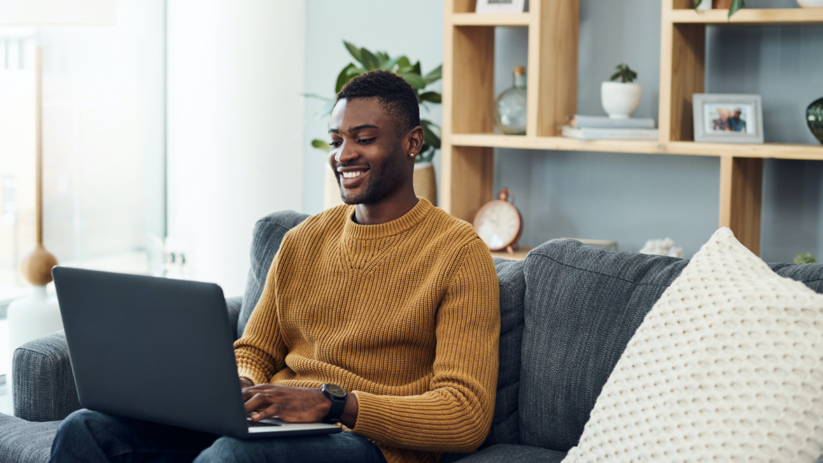 Ein junger Mann sitzt mit seinem Laptop auf einem Sofa und lächelt. (Quelle: iStock | Nicola Katie) Ein junger Mann sitzt mit seinem Laptop auf einem Sofa und lächelt.