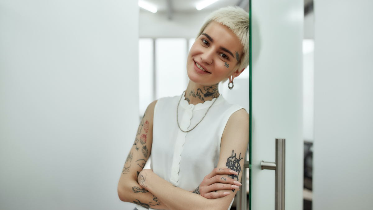 Eine junge Frau mit blonden Haaren und Tätowierungen lehnt sich an eine Glastür. (Źródło: iStock | LanaStock) Eine junge Frau mit blonden Haaren und Tätowierungen lehnt sich an eine Glastür.