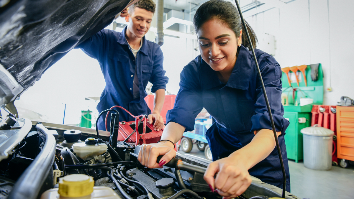 Eine junge Frau und ein junger Mann in Arbeitskleidung blicken in den Motorraum eines Autos. (Źródło: iStock | JohnnyGreig) Eine junge Frau und ein junger Mann in Arbeitskleidung blicken in den Motorraum eines Autos.